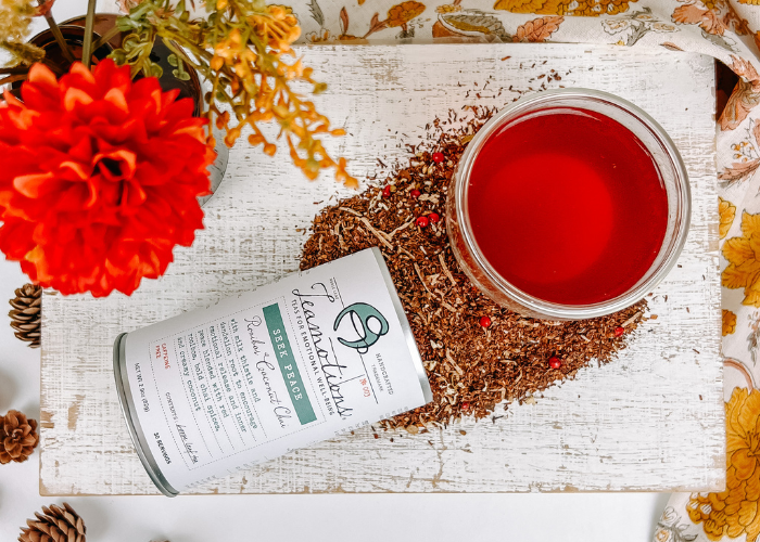 Red beverage in a glass, tea canister, and flowers on a white wooden surface.