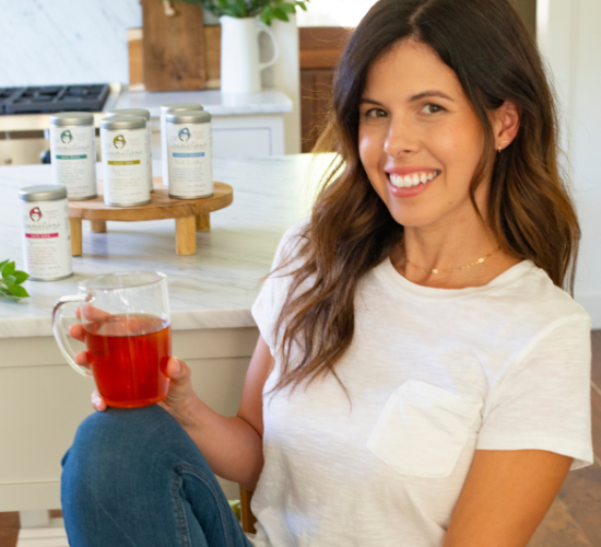 Woman smiling, holding a glass of red tea in a kitchen with tea canisters on the counter.
