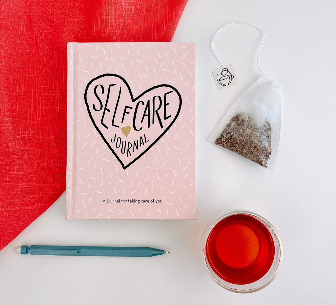 Self-care journal with a pencil, tea bag, and cup of tea on a white surface.