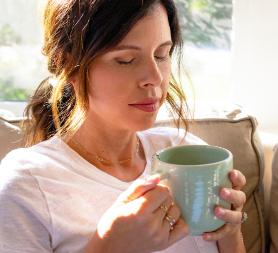 Person holding a green mug, eyes closed, sitting in a sunlit room.