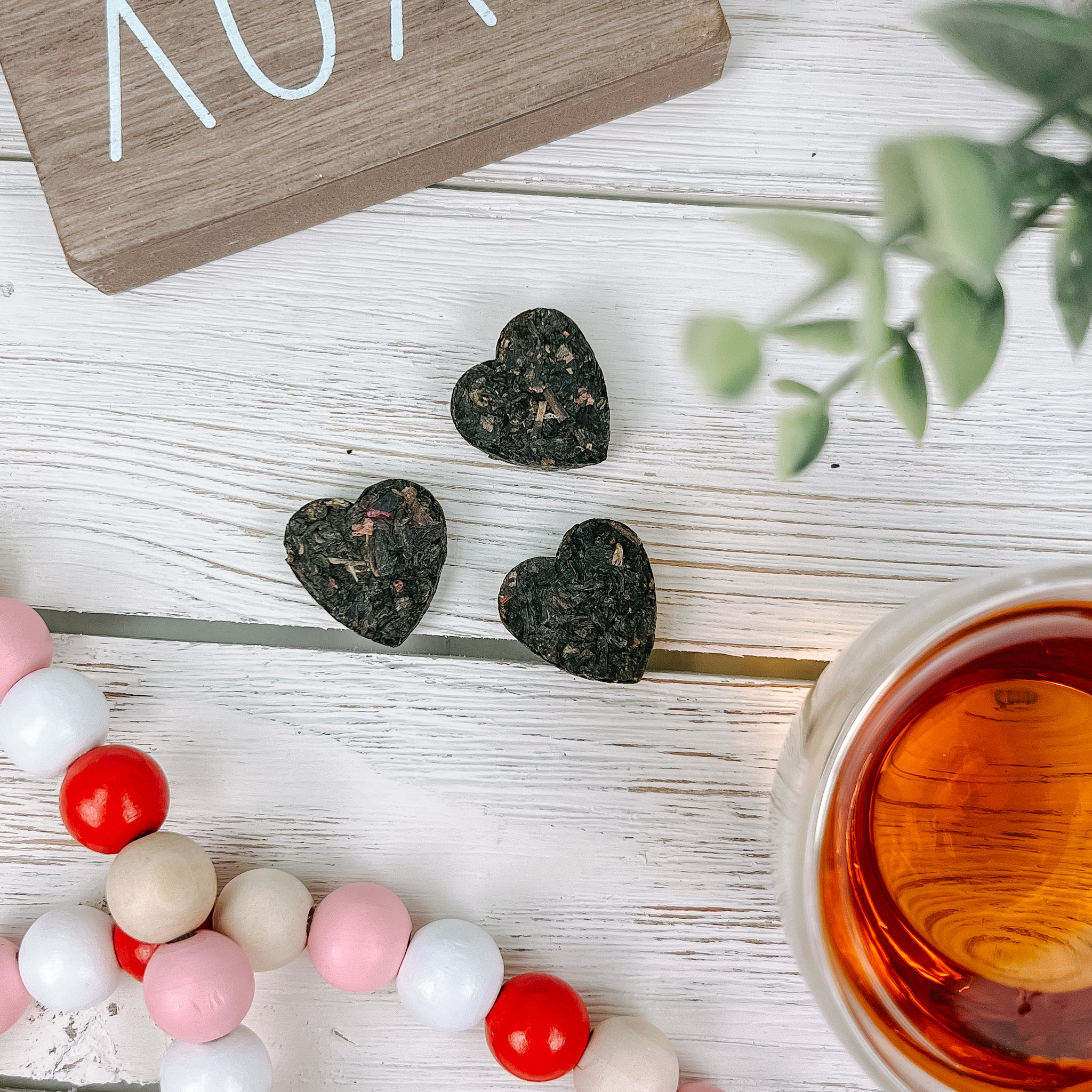 An overhead shot of heart-shaped pressed tea, a glass of tea, and colorful beads on a whitewashed table.