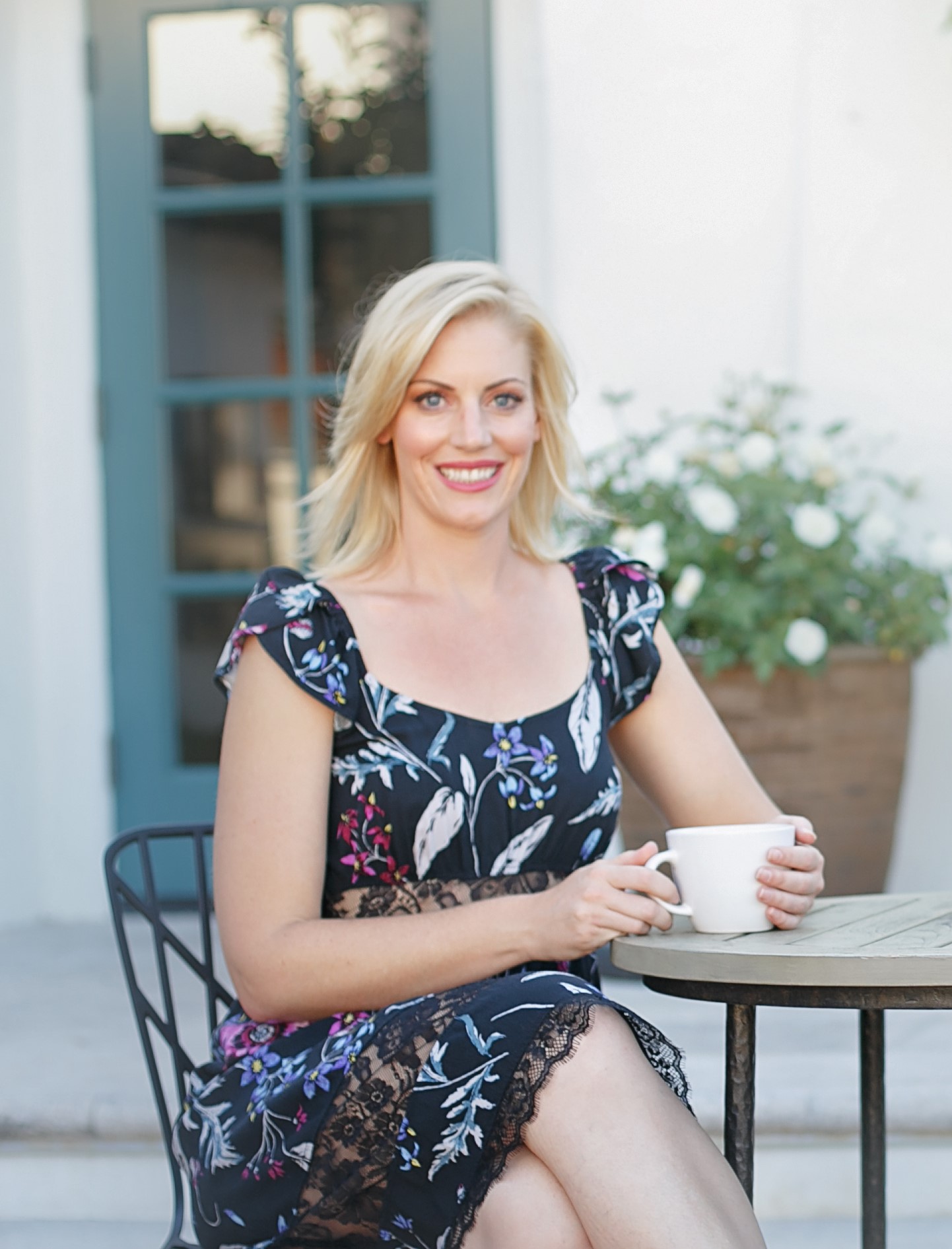 A blonde woman in a floral dress sits at an outdoor table, smiling and holding a white mug.