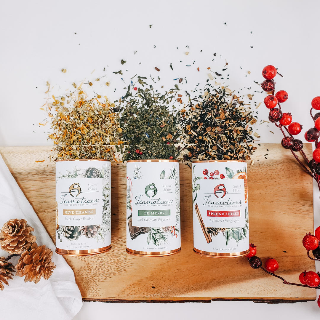 Three tea canisters with spilled tea leaves, pinecones, and red berries on a wooden surface.