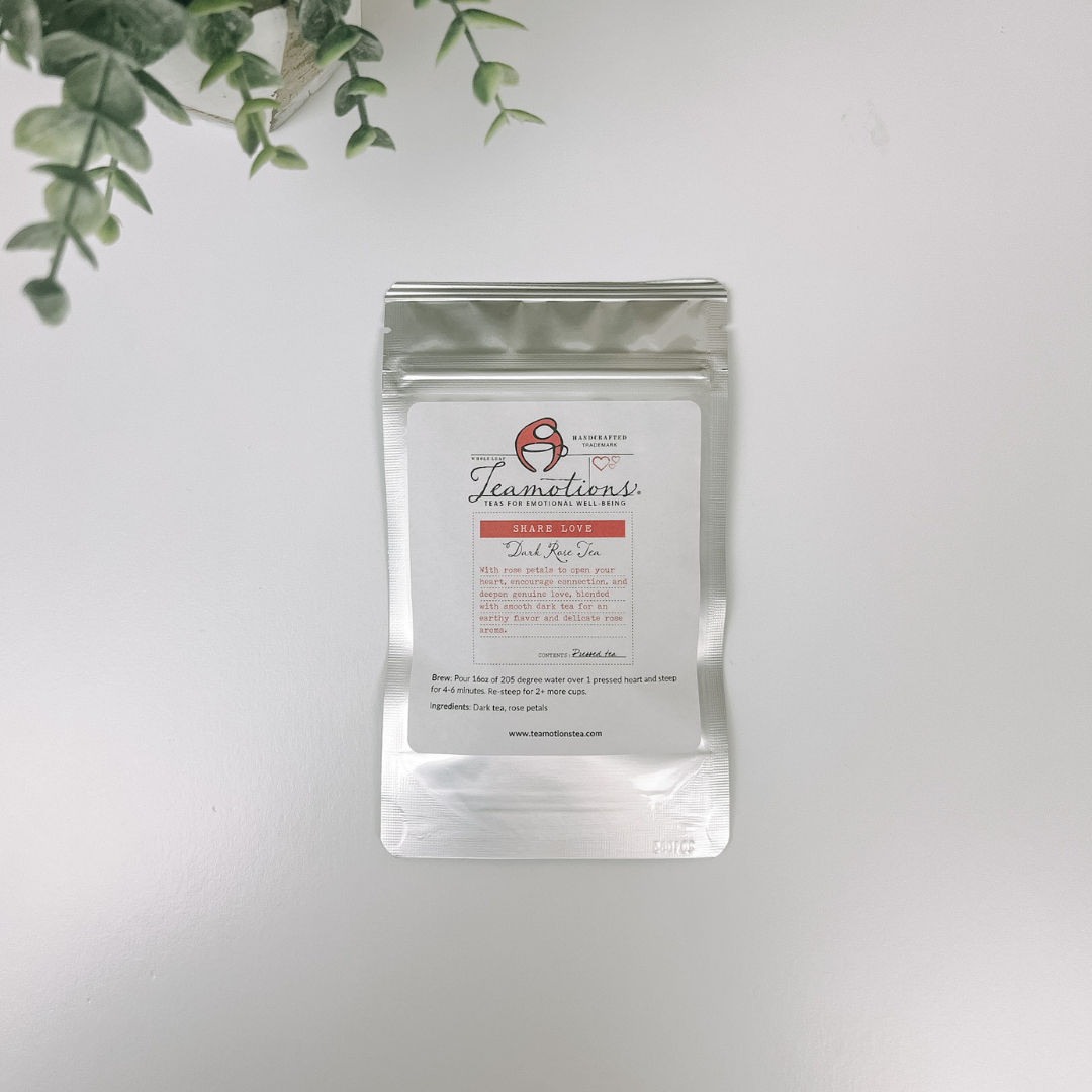 A silver pouch of Teamotions Dark Rose Tea on a white surface with a green plant.