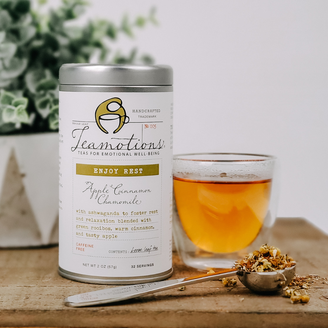 Tea tin labeled 'Teamotions,' glass of tea, scoop with loose tea, and plant in background.