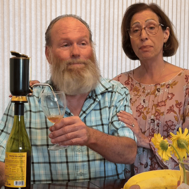 Two people with a bottle and glass at a table with flowers.