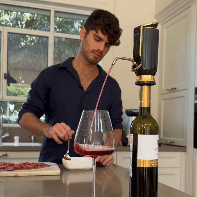 A man in a kitchen pours wine into a glass using a wine dispenser.