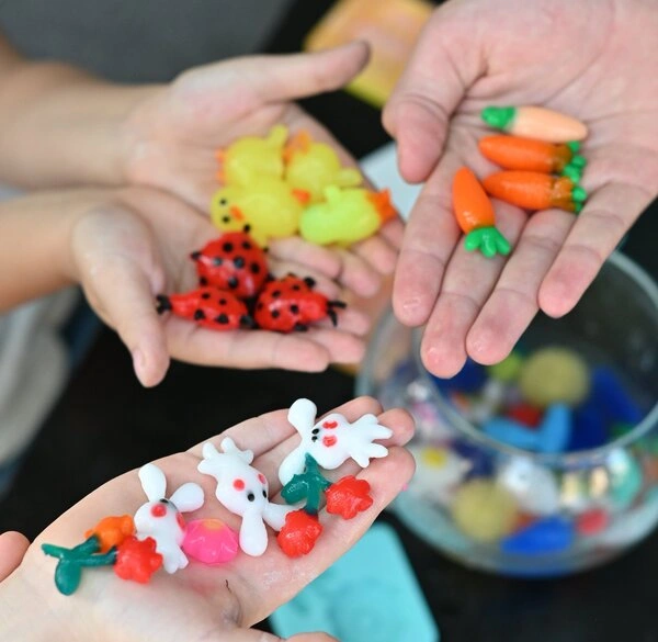 Hands holding small colorful plastic toys shaped like animals and vegetables.