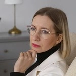 A woman with glasses, sitting and looking thoughtful in a room.