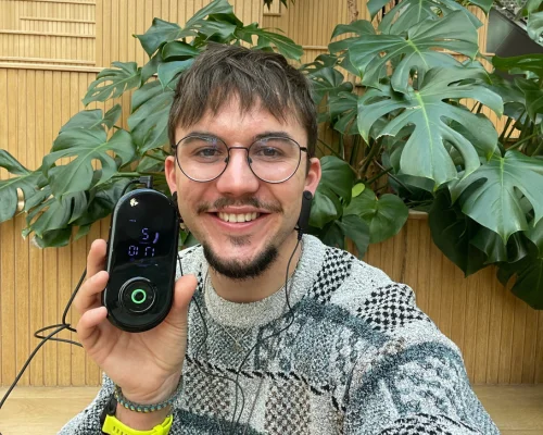 Man smiling, holding a portable device, in front of large green leaves.