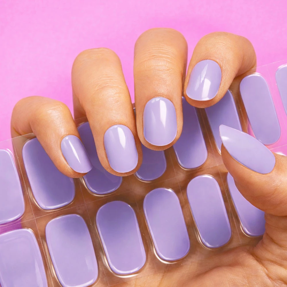 A hand with a lavender manicure holding a sheet of matching nail polish strips against a pink background.
