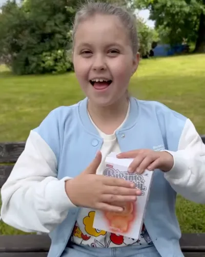 Child smiling and holding a book while sitting on a bench outdoors.