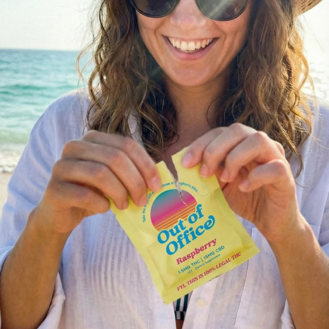 A smiling woman on a beach tears open a yellow packet that reads 'Out of Office'.