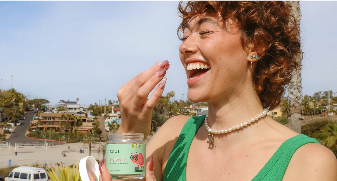 Person smiling, holding a jar labeled 'Soul Big Brain Focus Capsules' outdoors.