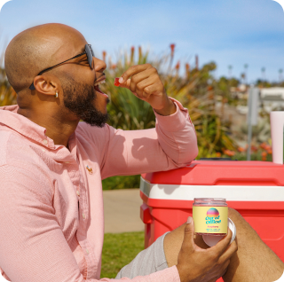 Man in sunglasses eating from a jar outdoors, sitting by a red cooler.
