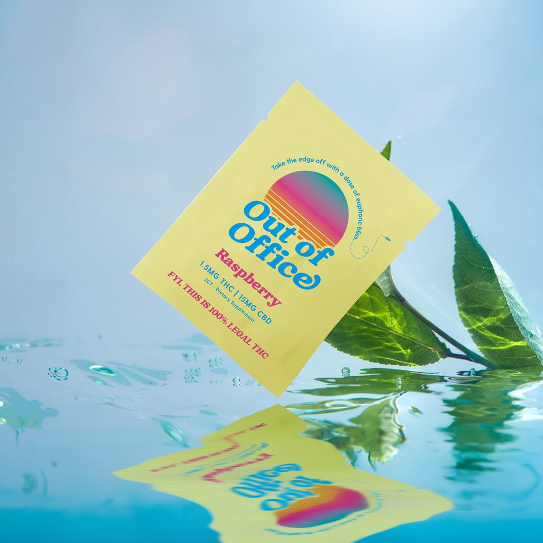 A yellow packet of Out of Office Raspberry supplement sits in water with its reflection and green leaves.