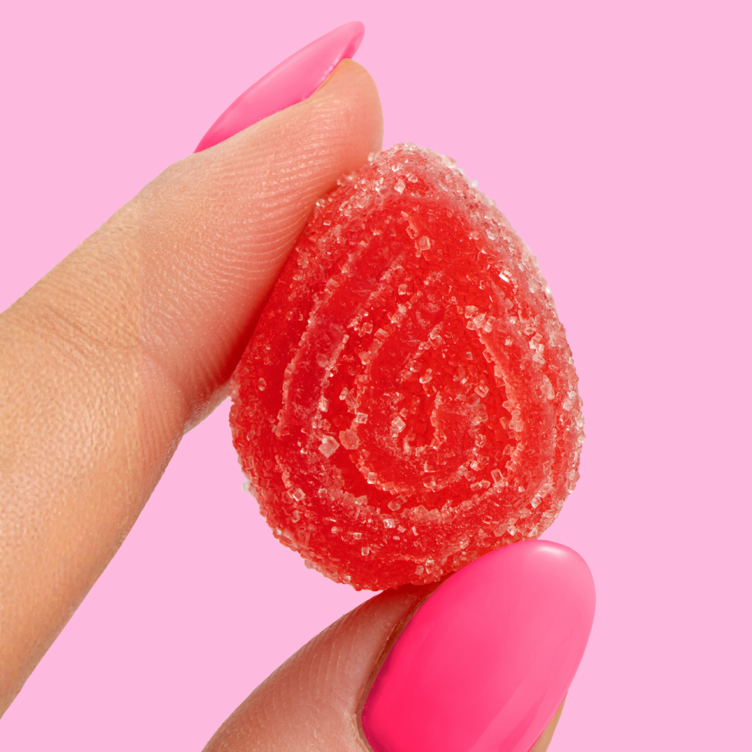 A close-up of a hand with pink nails holding a red, sugar-coated gummy against a pink background.