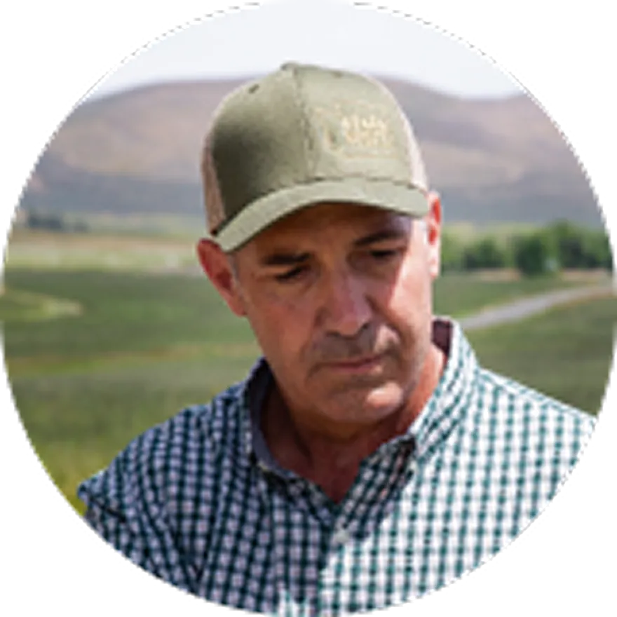 A circular portrait of a man in a green cap and plaid shirt, looking down outdoors.