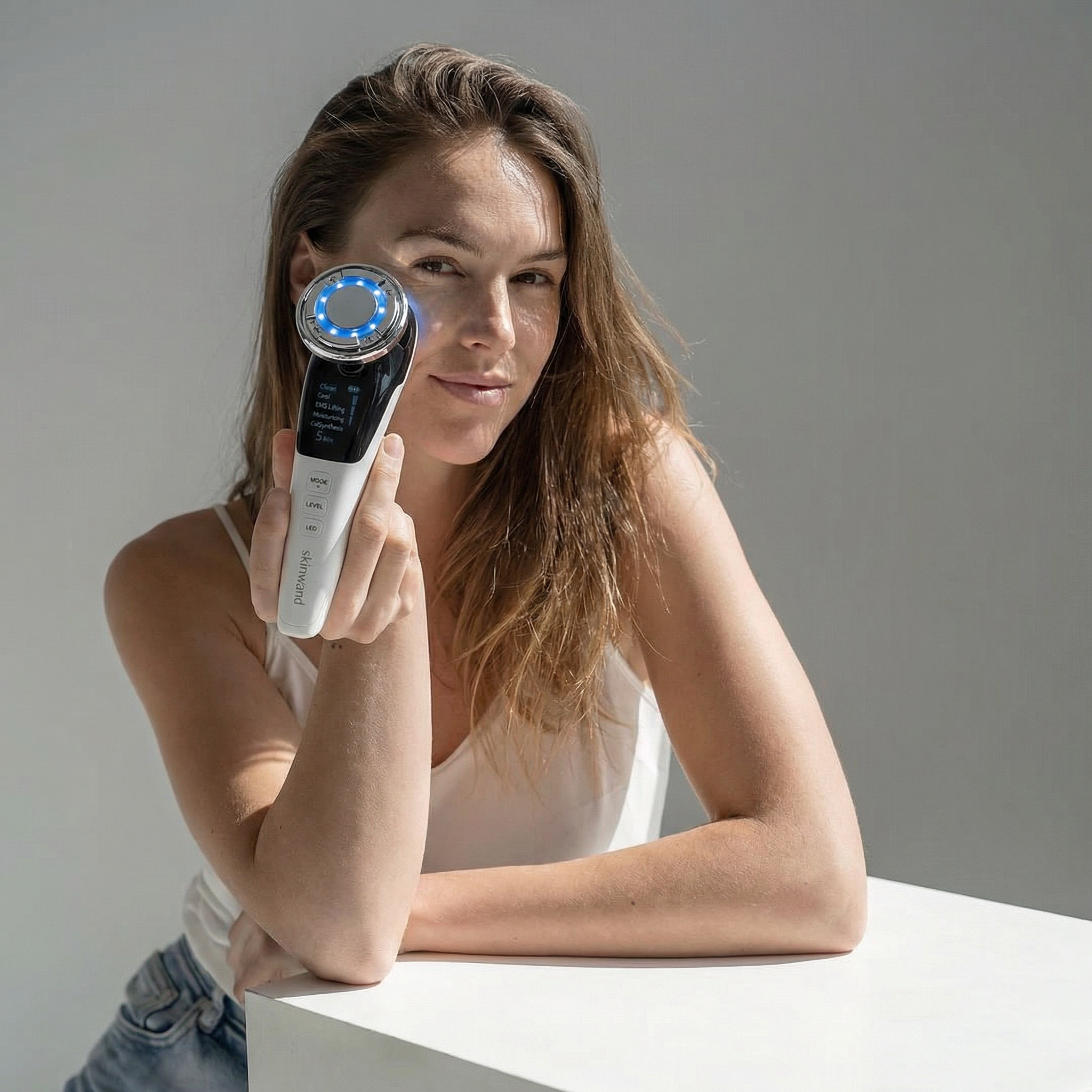 A woman holds an electronic skincare device with a blue light over one eye while looking at the camera.
