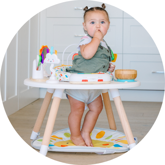 A baby girl with pigtails stands in a white activity center with toys around her.