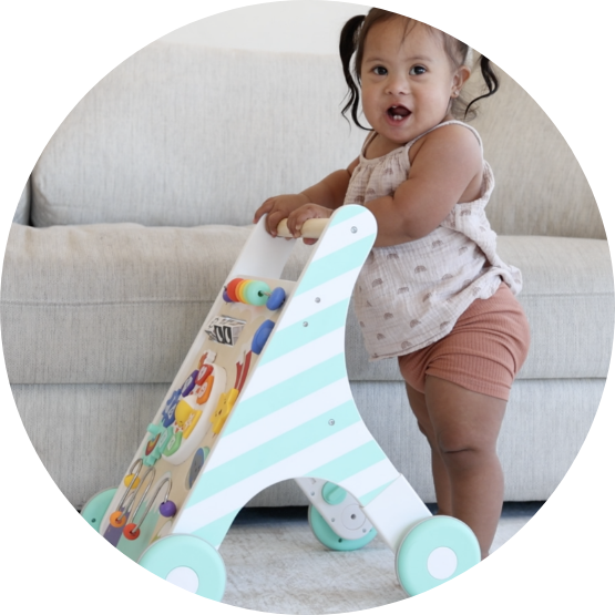 A smiling toddler with pigtails stands holding a wooden push walker with toys on it.