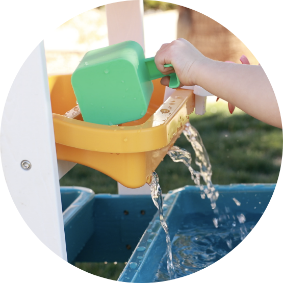 A child's hand uses a green scoop to pour water into a colorful plastic water table outdoors.