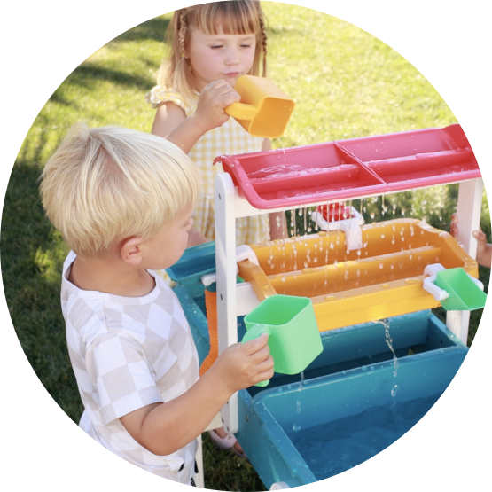 Two young children play with a colorful, multi-tiered water table outdoors on a grassy lawn.