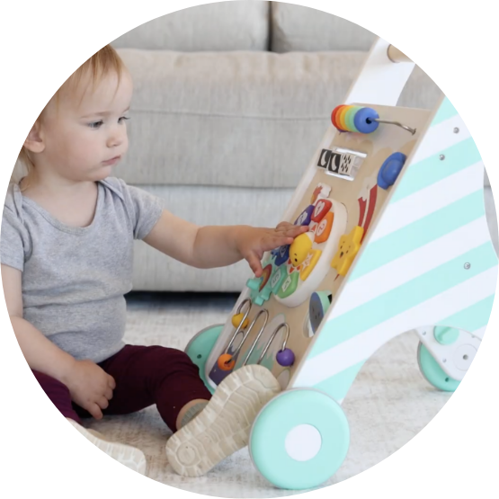 A toddler sits on the floor playing with a striped wooden activity walker toy.