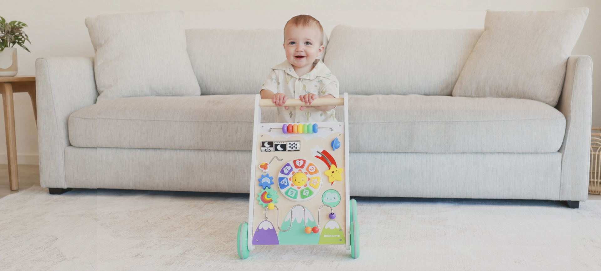 A smiling baby stands behind a colorful wooden activity walker in front of a light-colored couch.