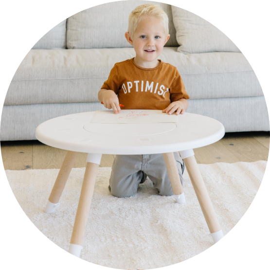 A smiling young boy kneels on a rug, drawing with a crayon at a small white table.