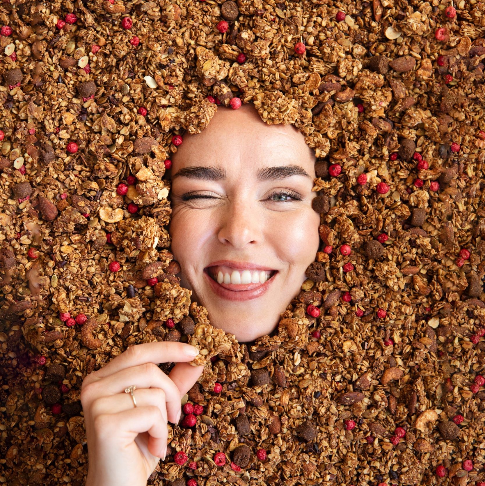 Smiling face surrounded by granola and berries, winking with hand holding granola.