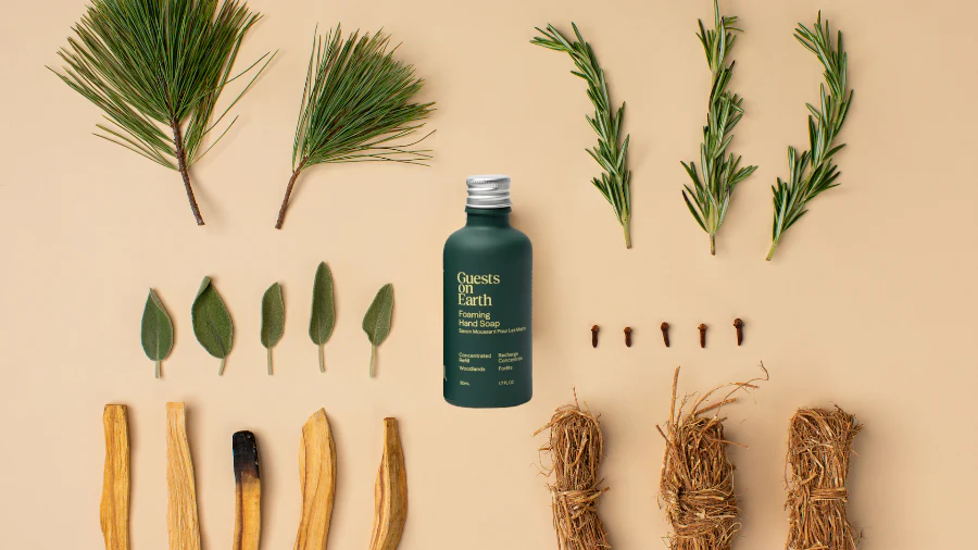 A bottle of foaming hand soap laid flat with botanicals including pine, rosemary, sage, cloves, and wood sticks.