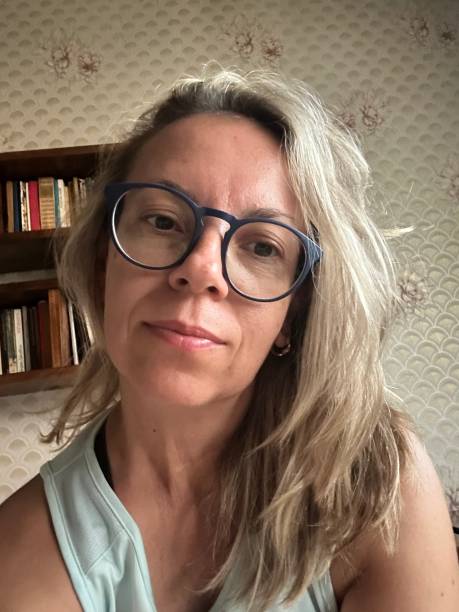 A woman with blonde hair and blue glasses looks at the camera in front of a bookshelf.
