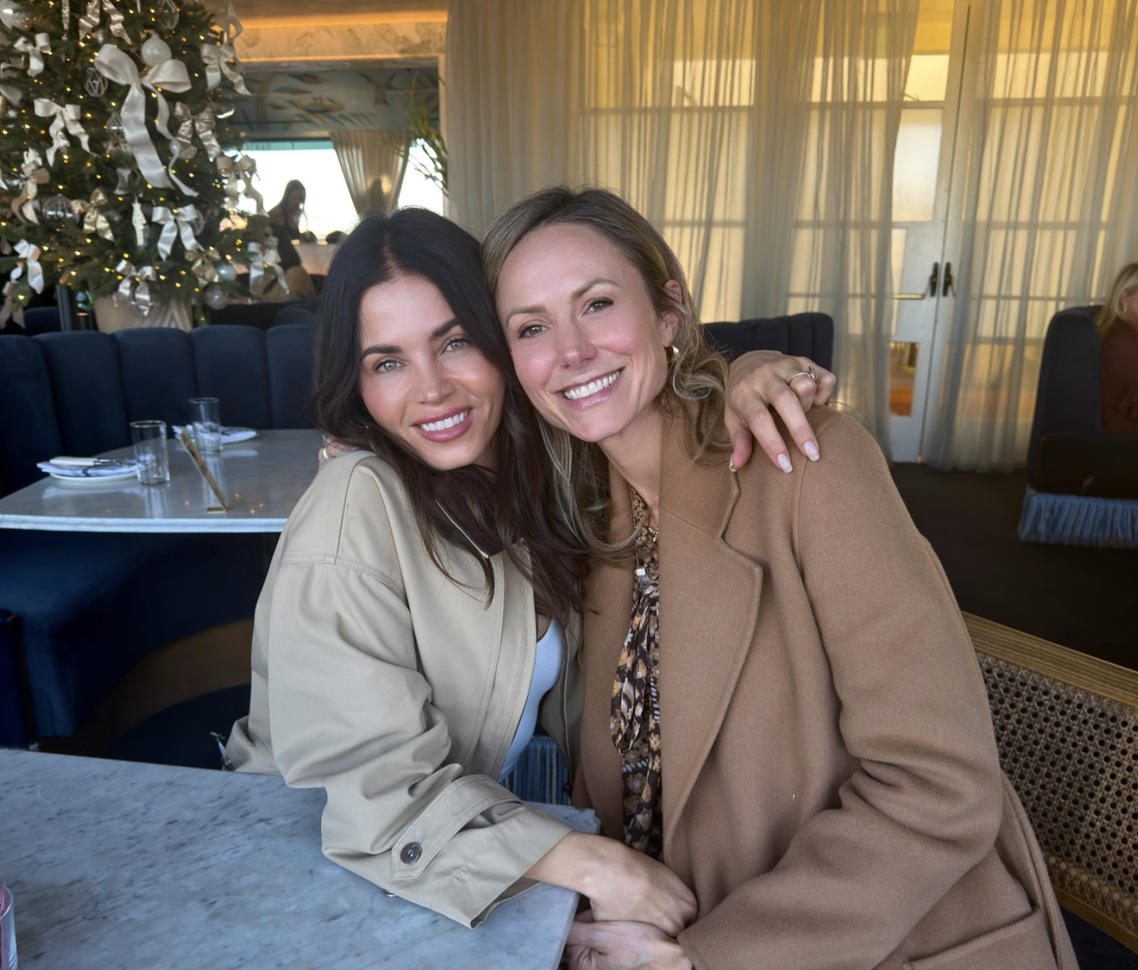 Two women smile and embrace while sitting at a table inside a restaurant.
