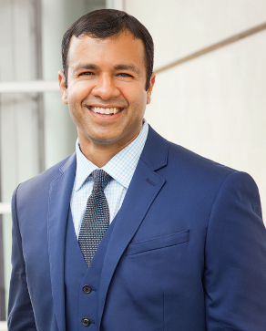 A headshot of a smiling man wearing a blue suit, vest, light blue shirt, and dark patterned tie.