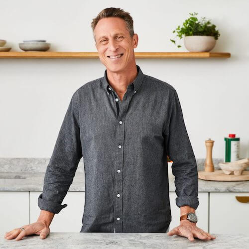 Smiling person standing in a kitchen with shelves and plants.