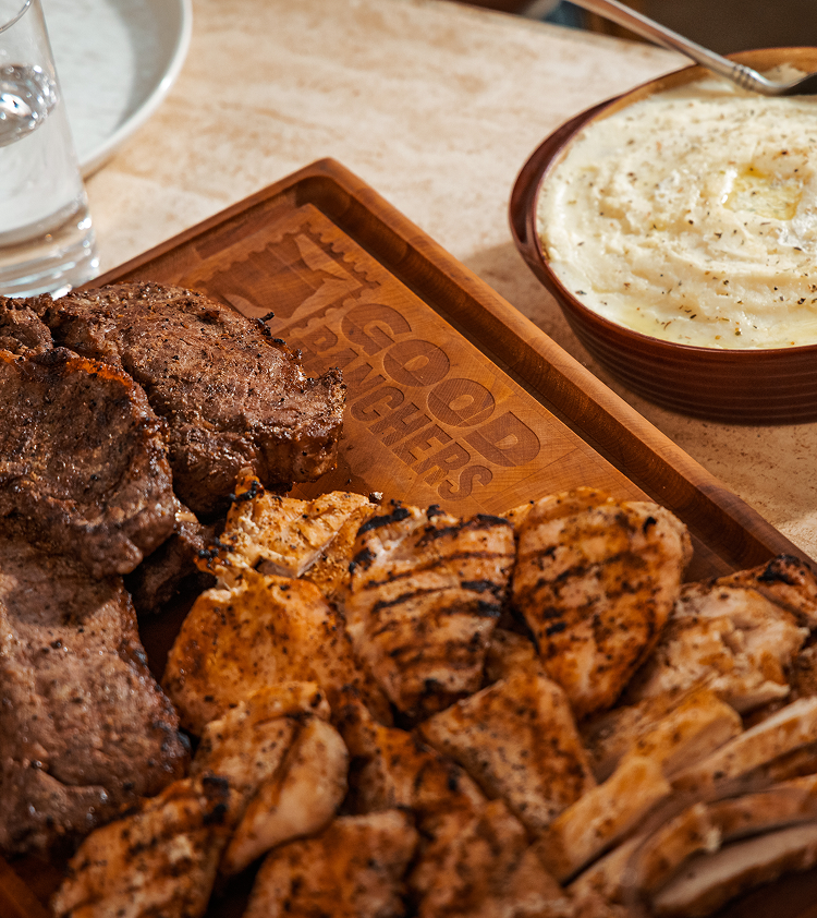 Grilled meats on a wooden tray next to a bowl of mashed potatoes.