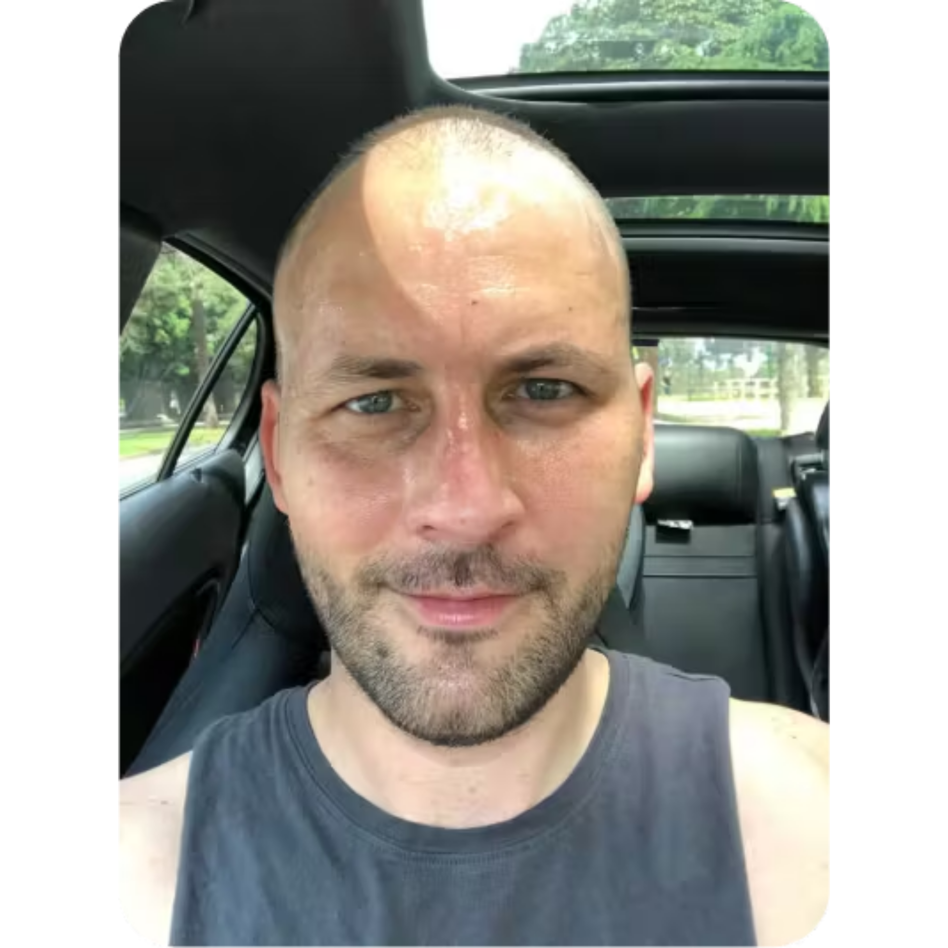 A person with a shaved head sitting in a car, wearing a sleeveless shirt.