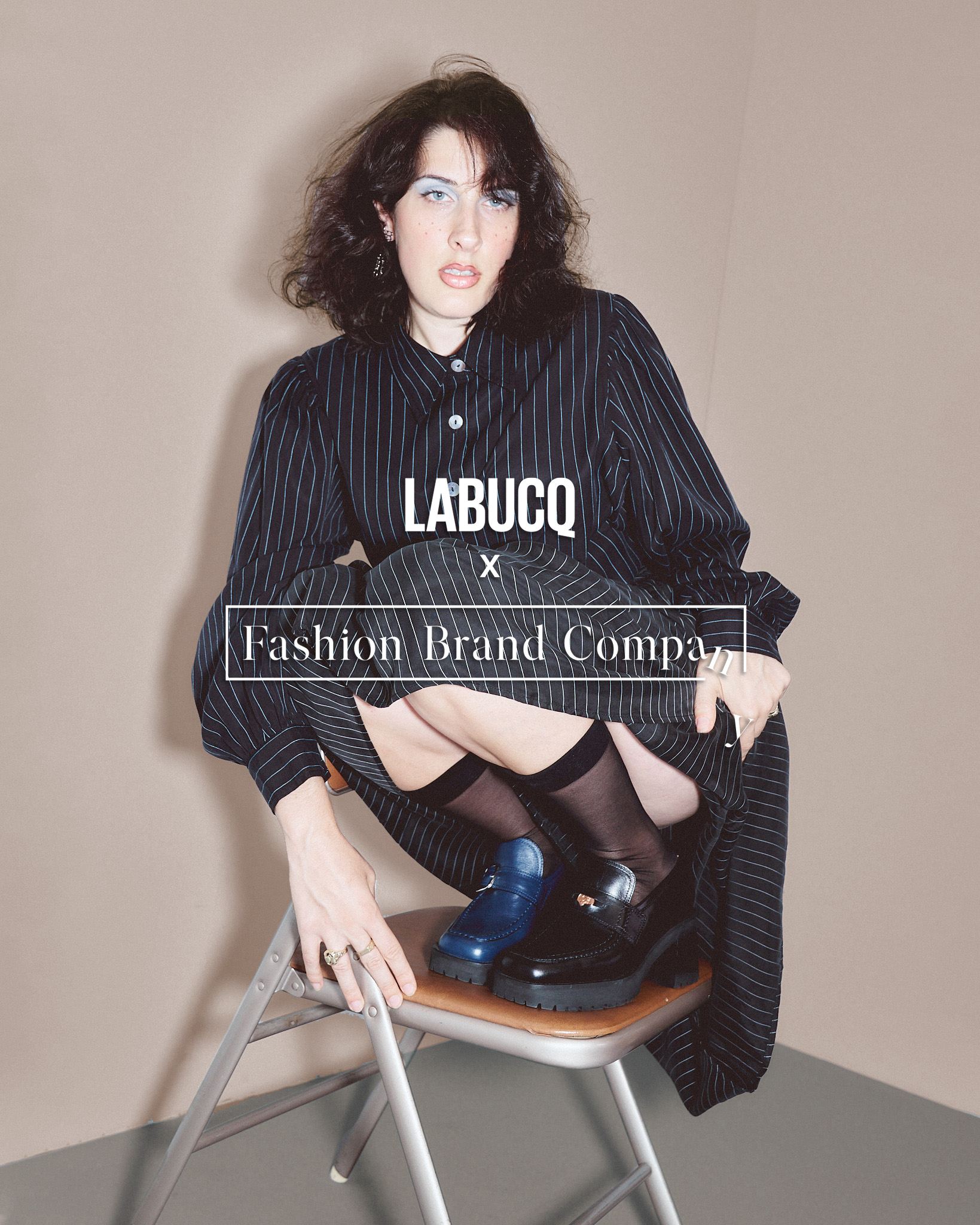 A model sitting on her heels atop a folding chair wearing a black striped dress and mismatched platform loafers. One loafer is a black penny loafer and the other is a dark blue loafer with a buckle.