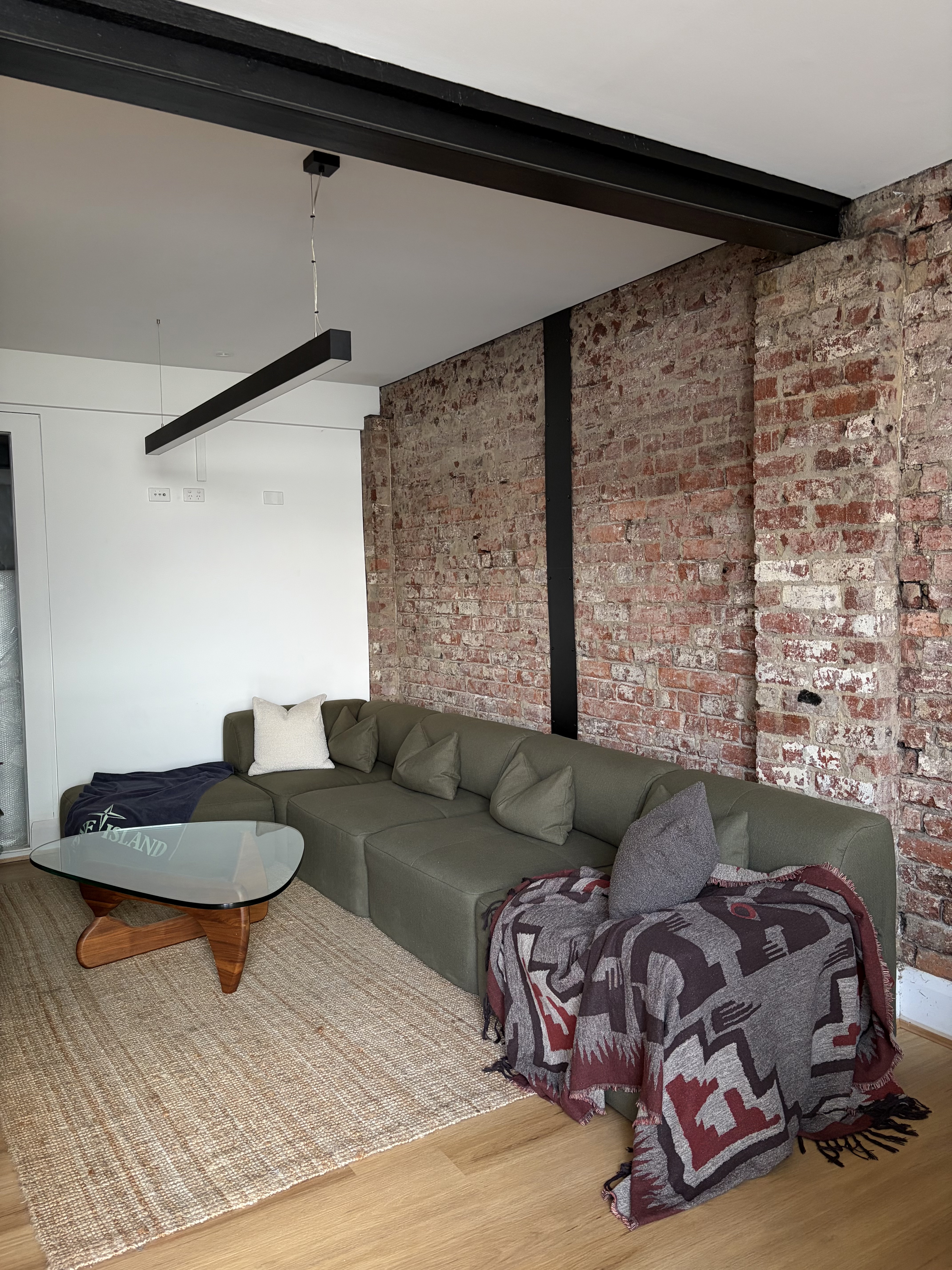 A living room with a green sectional sofa next to an exposed brick wall and a glass coffee table.