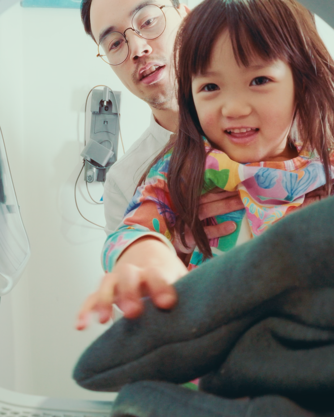 A man holds a young girl who is smiling and reaching towards laundry in a machine.