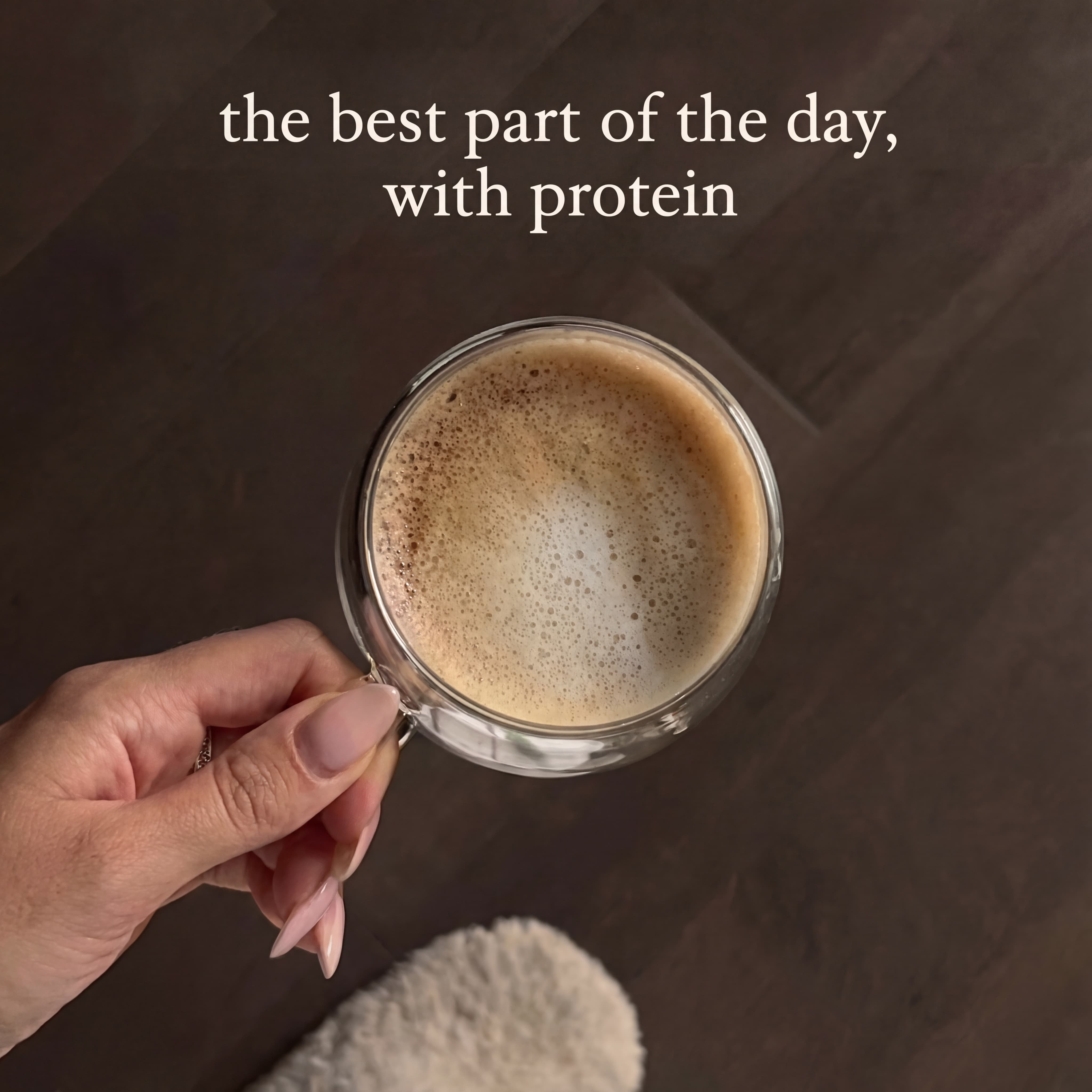 Hand holding a frothy coffee in a glass cup on a dark surface.