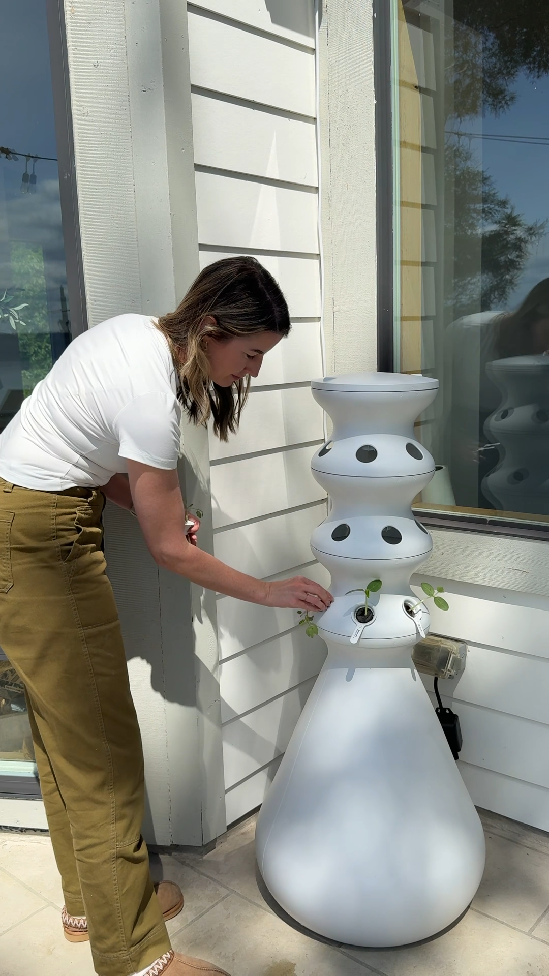A woman places seedlings into a white, tiered, vertical hydroponic garden tower on a patio.