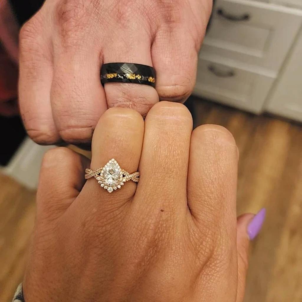 Two hands displaying a diamond engagement ring and a black band with gold accents.