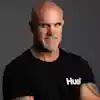 A person with a bald head and beard wearing a black shirt with 'Hustle' text.