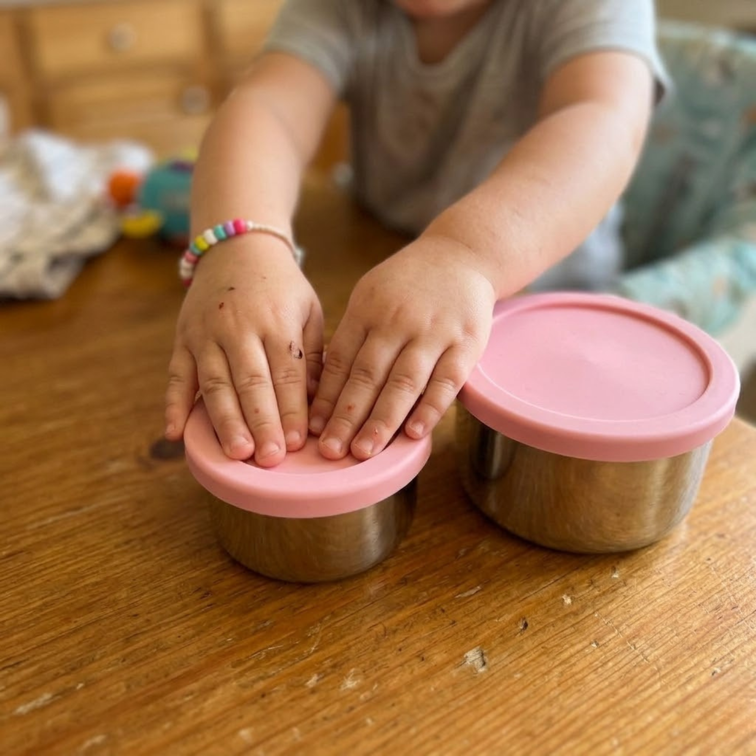 A toddler places their hands on top of two small stainless steel containers with pink lids.