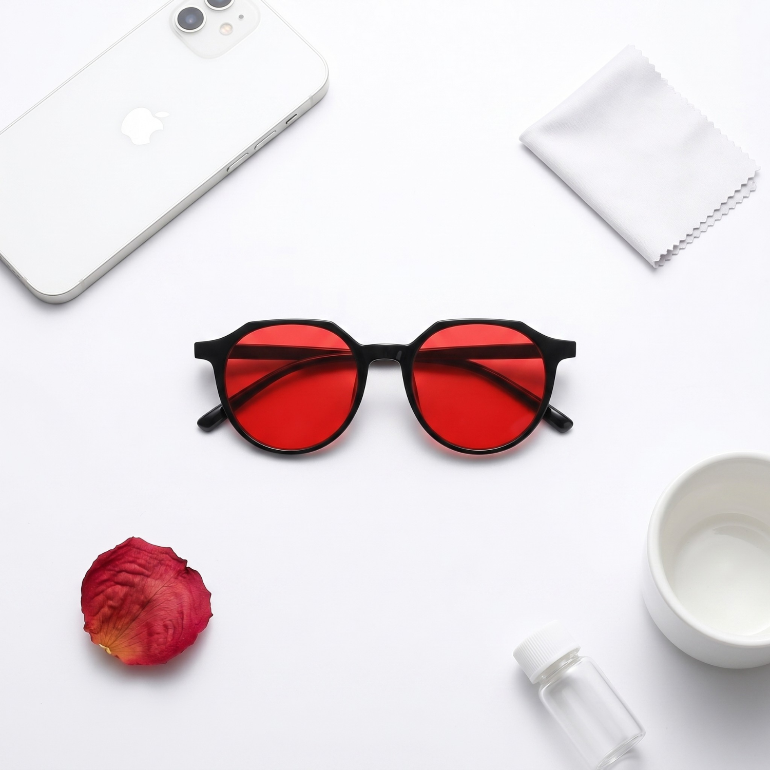 A flat lay of sunglasses with red lenses, a smartphone, a rose petal, and other accessories.