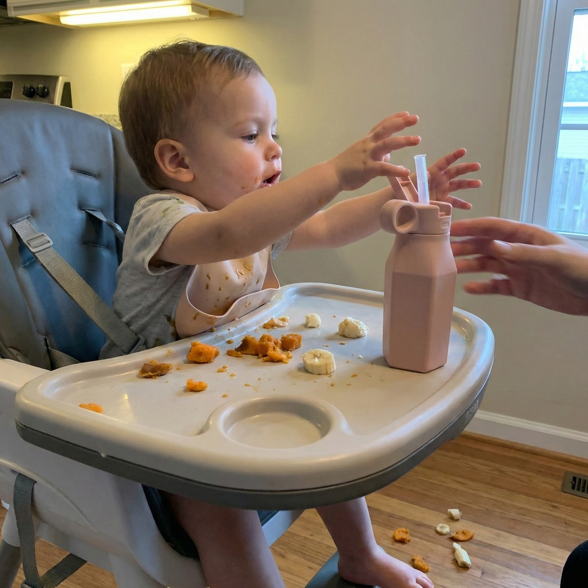 A baby in a high chair reaches for a pink bottle as an adult's hands steady it.