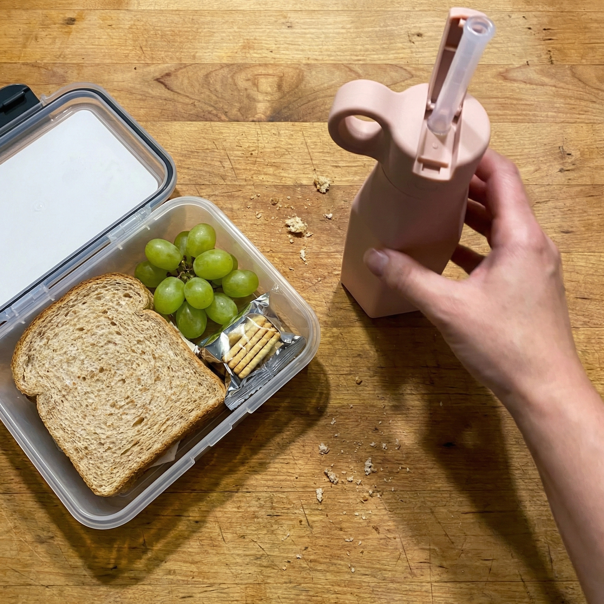 A person's hand holds a pink bottle next to an open lunch container with a sandwich and grapes.
