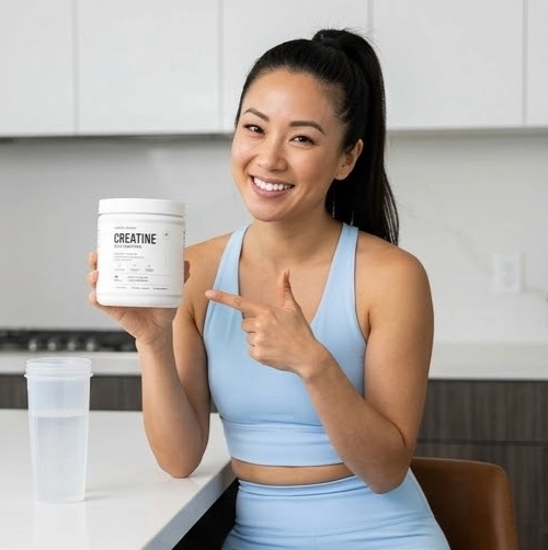A smiling woman in blue athletic wear sits at a counter holding and pointing to a jar of creatine.